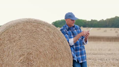 Farmer-using-digital-tablet--computer-while-standing-by-hay-bale.-Modertn-Agriculture