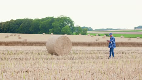 Farmer-using-digital-tablet--computer-while-standing-by-hay-bale.-Modertn-Agriculture