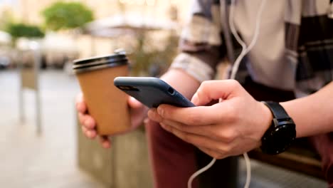 Man-in-headphones-using-smartphone,-listening-to-music-and-browsing-on-smartphone.-Drinking-coffee-to-go-outdoor.-Close-up.-Blogger-scrolls-through-social-media-on-device,-reading-news-on-app