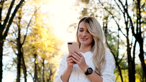 Retrato-de-chica-en-suéter-blanco-escribiendo-un-mensaje-en-su-teléfono-inteligente-al-aire-libre.-Mujer-usando-gadget-digital,-se-desplaza-a-través-de-las-redes-sociales-en-el-dispositivo,-leyendo-noticias-en-la-aplicación,-al-atardecer