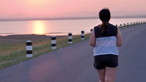 Übergewichtige-asiatische-Frauen-joggen-am-frühen-Morgen-im-Sonnenlicht-auf-der-Straße.-Konzept-des-Gewichtsverlustes-mit-Bewegung-für-die-Gesundheit.-Langsame-Bewegung,-Rückansicht