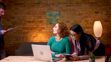 Closeup-Shooting-von-jungen-kaukasischen-Geschäftsfrauen,-die-am-Laptop-arbeiten-und-mit-zwei-Kollegen-über-Daten-diskutieren.-Mitarbeiterinnen-mit-Tablet