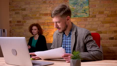 Closeup-shoot-of-adult-caucasian-businessman-working-on-the-laptop-indoors-in-the-office.-Businesswoman-handling-him-papers-sitting-the-couch-and-using-the-tablet-on-the-background