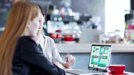 Zoom-out-of-two-young-businesswomen-discussing-website-templates-presented-on-laptop-screen-sitting-at-table-in-cafe,-Asian-woman-explaining-design-to-Caucasian-colleague