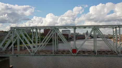 Vista-aérea-del-puente-ferroviario,-con-un-tren-en-movimiento-en-él,-a-través-del-río-que-fluye-a-través-de-una-ciudad-importante