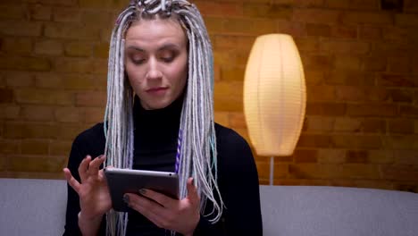 Closeup-shoot-of-young-attractive-caucasian-hipster-female-using-the-tablet-and-looking-at-camera-sitting-on-the-couch-indoors-in-a-cozy-apartment