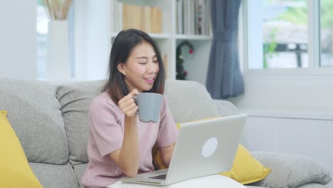 Young-business-freelance-Asian-woman-working-on-laptop-checking-social-media-and-drinking-coffee-while-lying-on-the-sofa-when-relax-in-living-room-at-home.-Lifestyle-women-at-house-concept.