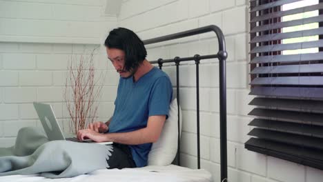 Young-Asian-man-using-laptop-checking-social-media-feeling-happy-smiling-while-lying-on-bed-after-wake-up-in-the-morning,-Attractive-male-smiling-relax-in-bedroom-at-home-concept.