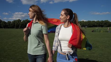 Novias-felices-con-bandera-lgbt-caminando-sobre-el-césped