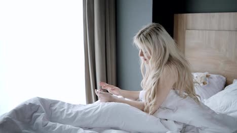 Use-the-phone.-Checks-email-and-social-media-messages.-Young-woman-wake-up,-sits-on-comfortable-bed.-Morning-in-hotel-room.