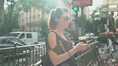 Young-attractive-woman-writing-a-text-message-on-her-smartphone-at-the-subway-exit-in-street,-drinking-her-coffee,-during-sunny-summer-in-Paris.-Freckles,-sunglasses,-piercings,-red-hair,-4K-UHD.