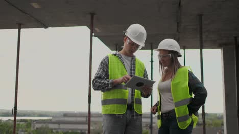 Trabajadorde-la-construcción-hombre-y-arquitecto-mujer-en-un-casco,-discutir-el-plan-de-construcción-de-la-casa,-hablarse-unos-a-otros-sobre-el-diseño,-sosteniendo-una-tableta,-mirar-los-dibujos,-fondo-de-los-rayos-del-sol