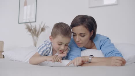 En-el-dormitorio-blanco,-mamá-e-hijo-miran-la-pantalla-de-la-tableta-y-se-ríen.-Familia-feliz-en-la-cama-por-la-mañana-leyendo-un-libro-y-haciendo-clic-en-el-panel-táctil-de-la-computadora