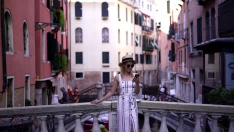 Efecto-de-cámara-lenta-de-chica-hipster-alegre-vestida-con-aspecto-casual-descansando-en-el-puente-de-Venecia-con-el-gadget-del-teléfono-inteligente-en-las-manos