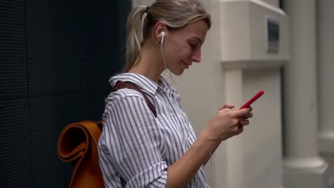 Positive-hipster-girl-enjoying-listening-playlist-via-modern-earphones-while-holding-new-mobile-phone-in-hand-and-reading-funny-news-in-blog