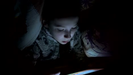 A-child,-a-little-girl,-hid-under-a-blanket-on-the-bed-at-night-and-plays-on-a-tablet-in-the-dark.-Concept-video.-Internet-addiction.-Close-up-of-the-face.-Raw-video.-4K.