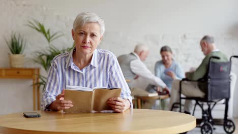 Rack-focus-of-three-elderly-people,-two-men-including-disabled-one-and-woman,-playing-cards-in-nursing-home.-Old-woman-with-short-grey-hair-reading-book-and-looking-at-camera-sitting-at-table