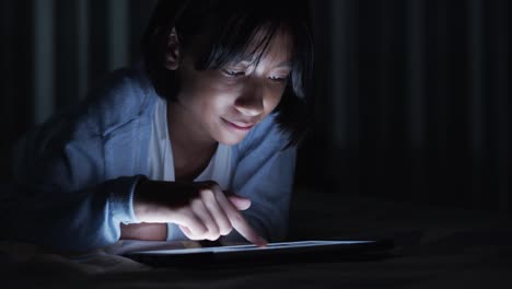 Asiatische-kleine-Mädchen-berührt-den-Bildschirm-auf-modernen-digitalen-Tablet-im-Schlafzimmer-in-der-Nacht.