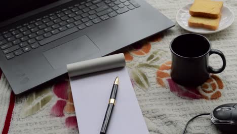 Breakfast-coffee-in-morning-sunlight-with-laptop-computer-black-color-pen-and-white-ruled-paper-notebook,-ceramic-cup-saucer-and-biscuit-on-top-office-place-working-desk-background.-Lifestyle-image.