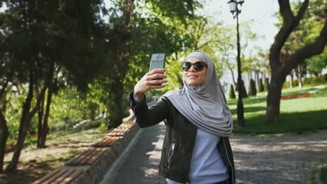 Muslim-female-in-casual-clothes,-sunglasses-and-hijab.-She-is-smiling,-enjoying-online-video-call-on-her-smartphone-while-walking-in-park.-Сlose-up