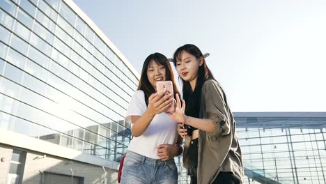 Beautiful-smiling-asian-girls-with-long-hair-and-dressed-in-stylish-clothes-standing-together-near-airport-and-using-their-phone