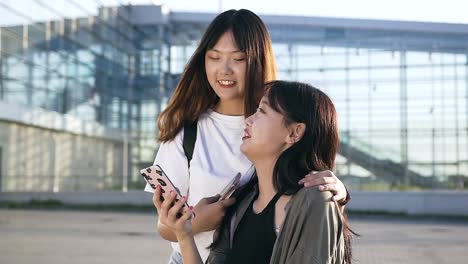 Dos-encantadoras-chicas-asiáticas-exuberantes-sonriendo-por-la-revisión-de-fotos-divertidas-en-el-teléfono-cerca-del-edificio-moderno