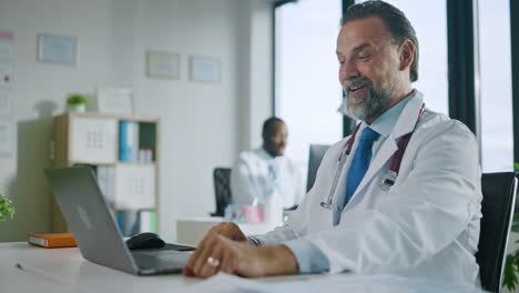 Hausarzt-macht-einen-Videoanruf-mit-Patient-auf-Laptop-Computer-in-einer-Gesundheitsklinik.-Physician-in-Lab-Coat-spricht-über-Gesundheitsprobleme-und-Diagnose-im-Krankenhausbüro.