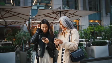 Asiatische-reife-Mutter-und-erwachsene-Tochter-benutzen-ihre-Smartphones-und-lachen,-während-sie-in-der-Nähe-der-Terrasse-des-Restaurants-stehen