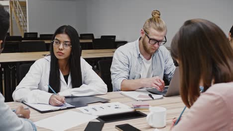 Unknown-boss-discussing-details-of-startup-project-with-young-team-of-diverse-colleagues,-sitting-at-table-in-modern-office