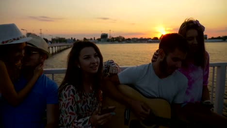 Group-of-friends-sitting-on-a-bench-and-singing-on-a-bridge-over-the-sea-in-slow-motion