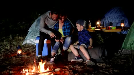 phone-in-hands-of-female-that-rests-with-husband-and-son-at-forest,-family-looking-photo-on-cell-phone-at-travel-camp