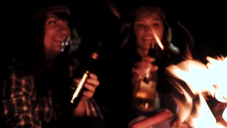 young-couple-drinking-beverage-and-doing-clink-glasses-near-fire-in-night-forest,-lovers-drink-alcohol-and-makes-toasting-glass-outdoors,-guy-with-girl-cheers-beer-in-travel-camp-at-woodland