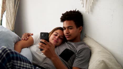 Happy-multiethnic-young-couple-relaxing-in-bedroom-with-the-smartphone-embracing-each-other.-Slowmotion-shot