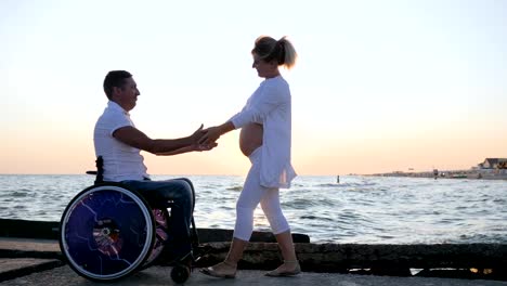 Pregnant-female-at-sunset-walks-up-to-disabled-man-in-a-wheelchair.-The-man-caresses-the-womans-pregnant-stomach-on-embankment