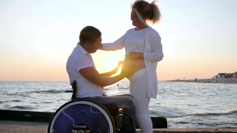 Love,-pregnant-female-with-disabled-husband-in-wheelchair-at-sunset