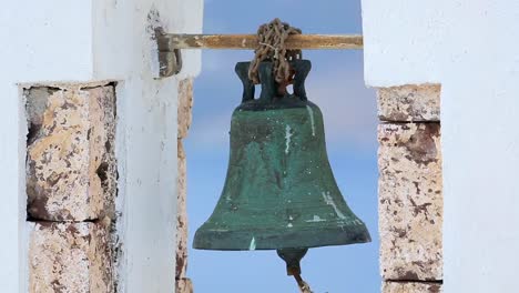 Old-rusty-bell-hanging-in-white-arch-against-blue-background,-church-in-Greece