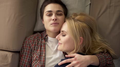 Two-young-lesbian-girls-lie-on-the-couch,-hug,-cuddle,-sleep,-girl-with-short-hair-looks-at-the-camera,-lgbt-family-concept,-happy,-top-shot-60-fps