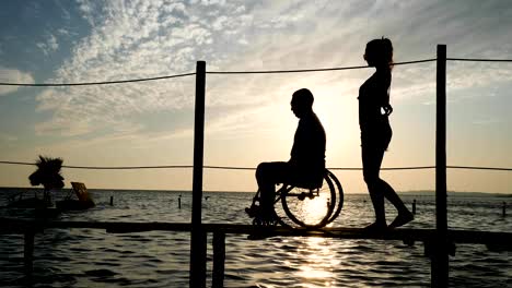 slender-girl-with-man-disabled-on-wheelchair-walking-on-pier-at-sea-against-sky-in-sunset