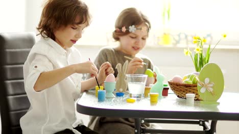 Hermano-y-hermana-pintan-huevos-de-Pascua-en-la-casa-en-la-cocina.-Están-concentrando.-Pinceles-y-pinturas,-creatividad-infantil