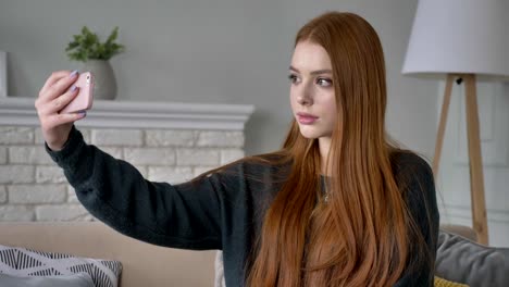 Young-beautiful-red-haired-teen-girl-uses-a-smartphone,-makes-selfie,-home-comfort-in-the-background.-60-fps