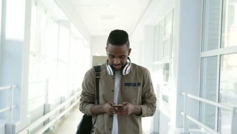 Handsome-afro-american-male-student-with-big-white-headphones-walking-in-long-lighty-corridor-of-college-holding-smartphone-texting-someone