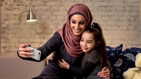 Junge-schöne-Mutter-Hijab-mit-kleinen-Mädchen-auf-der-Couch,-lächelnd,-nutzt-Smartphone-macht-Selfie,-Kuscheln,-kleines-Mädchen-mit-Teddybär,-glücklich-Familienkonzept-Wohnkomfort-im-Hintergrund-50-fps