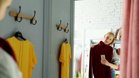 Female-customer-is-making-mirror-selfie-with-new-knitted-jumper-using-smart-phone.-She-is-standing-in-fitting-room-in-clothes-store-and-posing-with-lovely-smile.