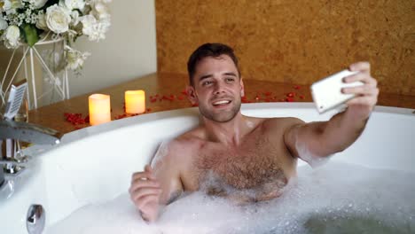 Junge-bärtigen-Kerl-berühmter-Blogger-ist-im-Whirlpool-im-DaySpa-mit-Smartphone-Videoaufnahme.-Brennende-Kerzen,-Glas-Champagner-und-Blumen-sind-sichtbar.