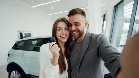 Point-of-view-shot-of-young-couple-have-online-vidoe-call-in-car-dealership-after-purchasing-car.-Man-is-holding-camera-and-woman-is-showing-keys,-they-are-talking-and-kissing.