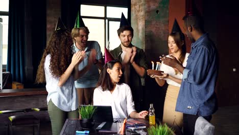 Mujer-bonita-está-trabajando-en-la-oficina-moderna-cuando-sus-colegas-están-llevando-el-pastel-de-cumpleaños-y-sombreros-del-partido,-chica-es-soplar-velas-y-palmas-de-las-manos-disfrutando-de-felicitaciones.