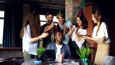 Group-of-young-people-is-congratulating-African-American-man-on-birthday-in-office-bringing-cake-and-clapping-hands,-guy-is-blowing-candles-and-having-fun.