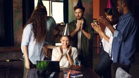 Attractive-young-woman-is-blowing-candles-on-birthday-cake-and-laughing-while-her-coworkers-are-clapping-hands-and-congratulating-her.-Party-and-work-concept.