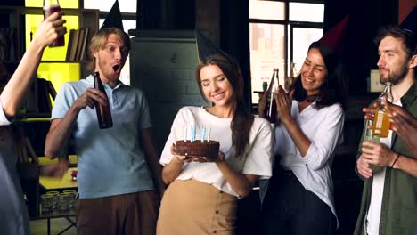 Company-employees-are-celebrating-birthday,-woman-is-holding-cake-and-blowing-candles,-her-coworkers-are-clapping-hands-and-clinking-bottles-with-drinks.