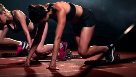 Tres-chicas-deportivas-en-ropa-de-color-negro-del-atleta-en-la-noche-en-la-cinta-se-iniciará-para-la-carrera-en-la-distancia-sprint-desde-la-posición-sentada